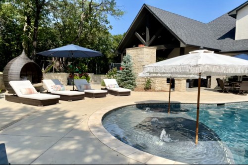 A sunny backyard with a pool, lounge chairs, umbrellas, and a modern home in the background surrounded by trees.