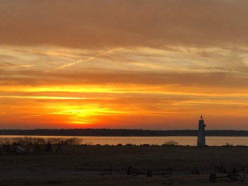 A serene sunset over a calm body of water, with a lighthouse silhouetted against vibrant orange and yellow skies.
