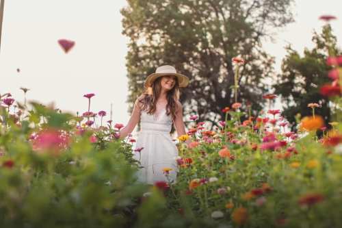 A woman in a white dress and straw hat stands among colorful flowers in a vibrant garden.
