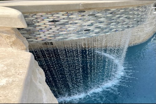 A close-up of a waterfall cascading over a stone edge into a blue pool, with shimmering tiles reflecting light.