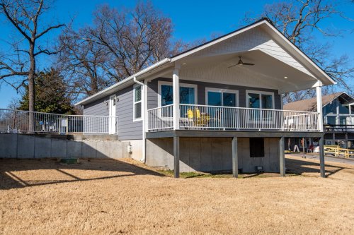 A modern gray house with a porch, surrounded by trees and a grassy yard, under a clear blue sky.