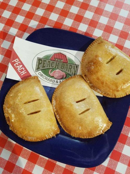 Three golden peach hand pies on a blue plate, with a logo from The Peach Barn on a red checkered tablecloth.