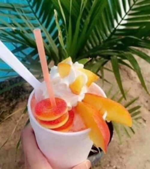 A refreshing fruit smoothie topped with whipped cream and peach slices, held in a white cup against a tropical backdrop.