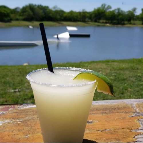 A refreshing margarita with a lime wedge sits on a wooden table, overlooking a serene lake and green landscape.