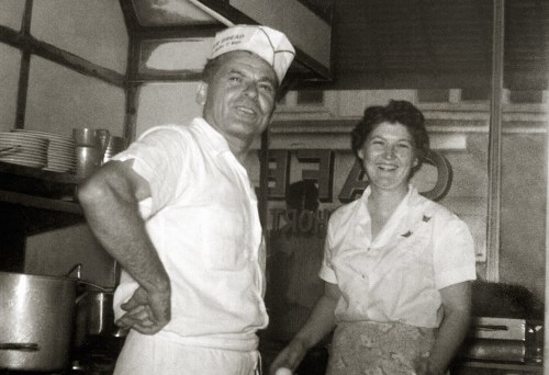 A smiling man in a chef's hat and a woman in an apron stand together in a vintage café kitchen.
