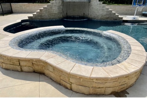 A curved hot tub with stone walls, surrounded by a pool and a waterfall feature in a sunny outdoor setting.