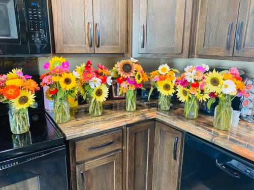 A kitchen countertop with seven vases of colorful flowers, including sunflowers and various blooms, arranged neatly.