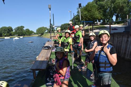 A group of kids in helmets and life vests smiles by a lake, preparing for a water sports activity.