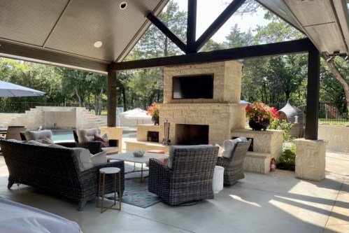 Cozy outdoor seating area with a stone fireplace, TV, and lush greenery in a serene backyard setting.