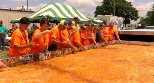 A group of people in orange shirts prepare a large dish at an outdoor event under a striped tent.
