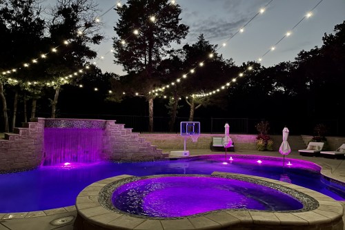 A beautifully lit pool area at dusk, featuring a waterfall, purple lights, and string lights overhead.
