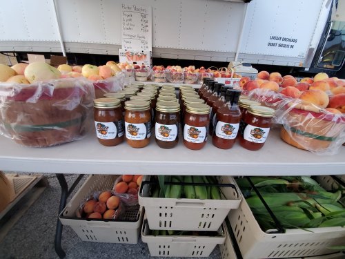 A table at a market displaying jars of preserves, baskets of peaches, and fresh produce.