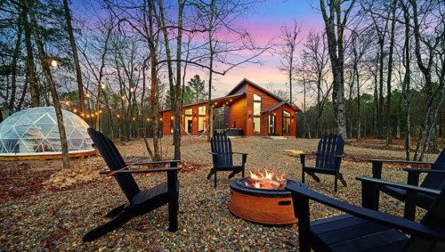 A cozy cabin surrounded by trees, with a fire pit and seating, and a geodesic dome in the background at sunset.