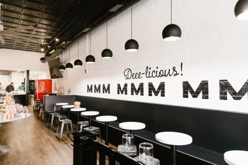 Interior of a modern eatery with a "Deee-licious! MMM MMM MMM" mural, black pendant lights, and white tables.