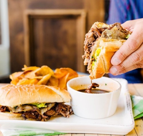 A hand holds a sandwich filled with meat and toppings, next to a bowl of dipping sauce and a plate of chips.