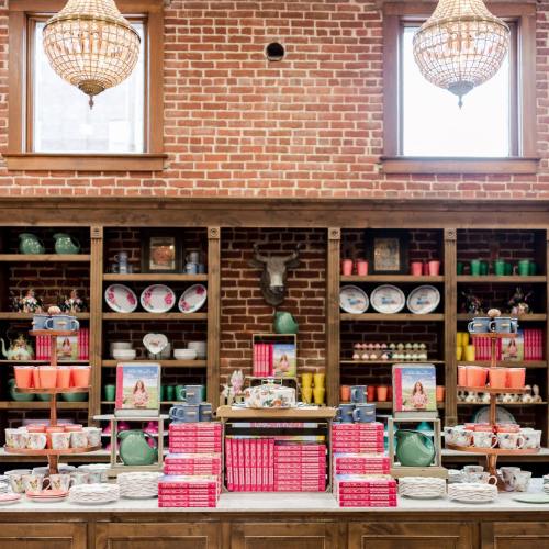 A charming display of colorful tableware and cookbooks on wooden shelves against a brick wall, with decorative lighting.
