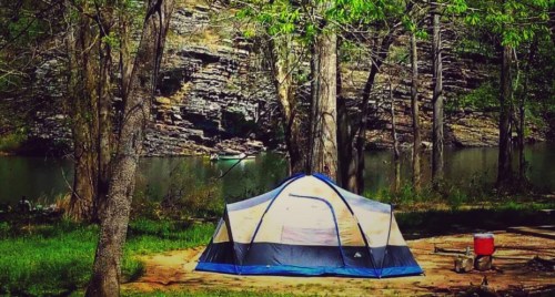 A colorful tent set up by a river, surrounded by trees and rocky cliffs in a serene outdoor setting.