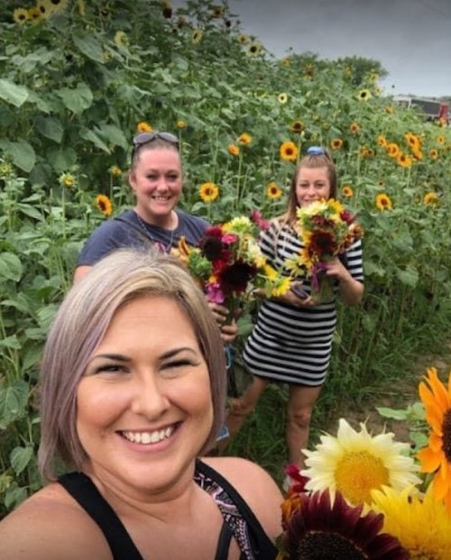 Three women smiling and holding flower bouquets in a sunflower field, surrounded by tall sunflowers.