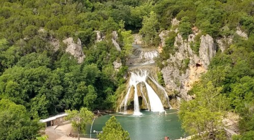 A scenic waterfall cascading into a green pool, surrounded by lush trees and rocky cliffs.
