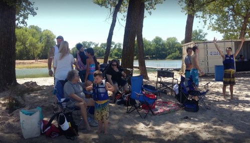 A group of people enjoying a sunny day at the beach, sitting in chairs near the water, with trees in the background.