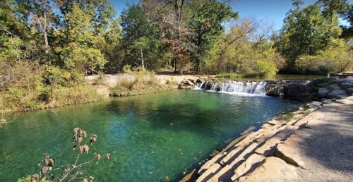 A serene natural scene featuring a clear green pond and a small waterfall, surrounded by lush trees and foliage.