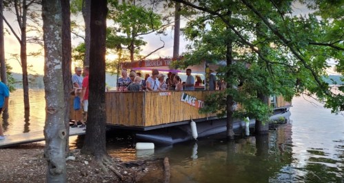 A wooden boat docked by the shore, with people gathered on it, surrounded by trees and a lake at sunset.
