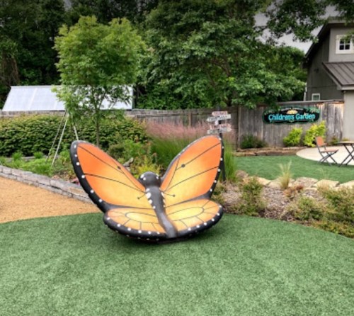 A large butterfly sculpture in a garden, surrounded by greenery and a sign for the Children's Garden.