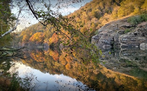 A serene river reflects colorful autumn foliage and rocky cliffs, framed by branches and a calm water surface.