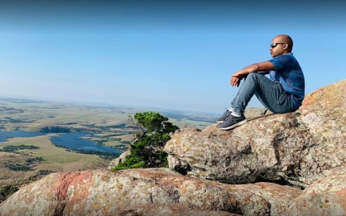 A person sitting on a rocky outcrop, overlooking a scenic landscape with lakes and rolling hills in the distance.