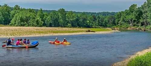 A group of people kayaking and rafting on a calm river surrounded by lush green trees and a sandy shore.