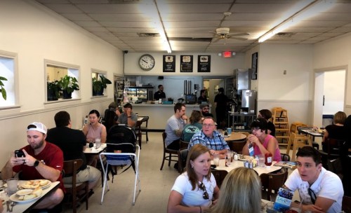A busy café interior with patrons dining, chatting, and using phones, featuring plants and a kitchen in the background.
