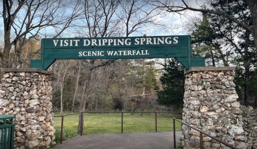Sign at the entrance to Dripping Springs Scenic Waterfall, framed by stone pillars and surrounded by trees.
