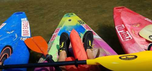 Three colorful kayaks on water, with a person's legs and paddles visible in the foreground.