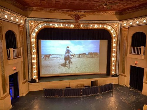 Interior of a vintage theater with a large screen showing a cowboy on horseback, ornate decor, and dim lighting.