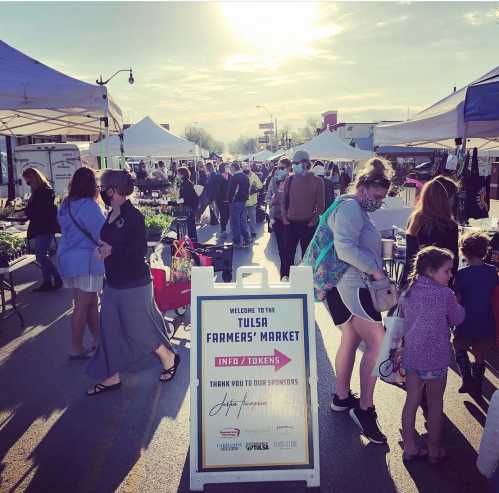 A bustling farmers' market with people browsing stalls, a sign welcoming visitors, and tents under a sunny sky.