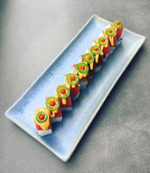 A platter of colorful sushi rolls topped with avocado, jalapeño, and drizzled with sauce, arranged neatly.