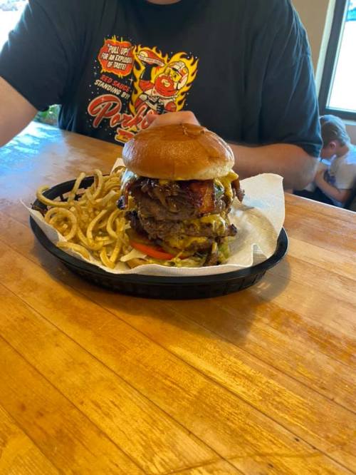 A large burger with bacon, cheese, and toppings, served with curly fries on a wooden table.