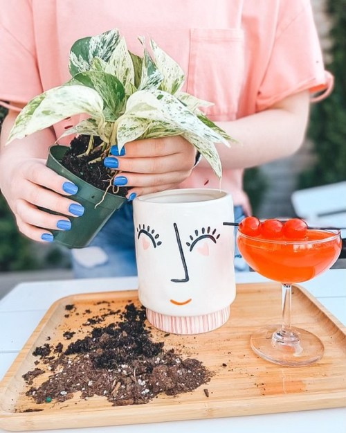 A person with blue nails holds a potted plant, preparing to repot it next to a cocktail with cherries on a wooden tray.