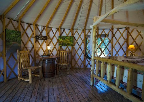 Cozy interior of a yurt featuring wooden furniture, a bed, and artwork on the walls. Warm lighting creates a welcoming atmosphere.