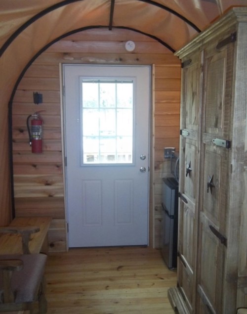 Interior of a cozy cabin with wooden walls, a door, a small fridge, and a wardrobe, featuring rustic decor.