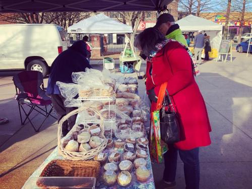 A woman in a red coat examines baked goods at a market stall, with tents and vendors in the background.