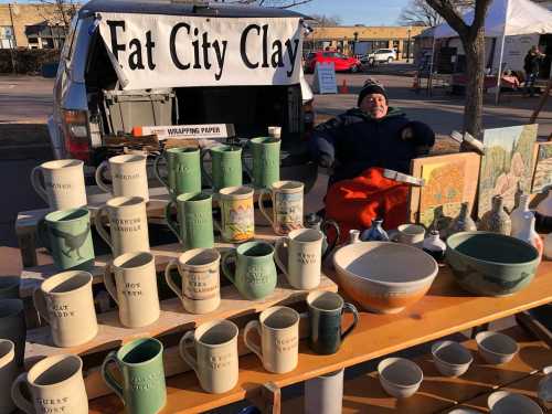 A vendor sits behind a table of pottery and mugs at a market, with a sign reading "Fat City Clay" in the background.