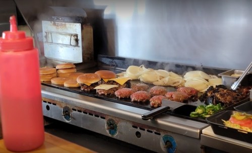 A grill with sizzling burgers, cheese, and onions, alongside bottles of condiments in a busy kitchen setting.