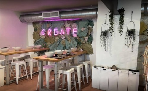 A modern dining space with a neon "CREATE" sign, wooden tables, white stools, and greenery on the walls.