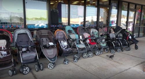 A row of various strollers displayed outside a store, with a bird walking on the pavement nearby.