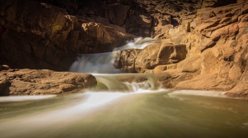 A serene waterfall cascades over rocky formations into a calm pool, surrounded by rugged terrain.
