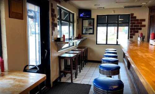 Interior of a casual diner with a wooden bar, blue stools, and a small table by the window. A TV is mounted on the wall.