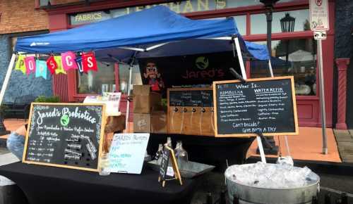 A market stall with a blue canopy, featuring signs for "Jared's Probiotics" and various beverages, with a colorful banner.