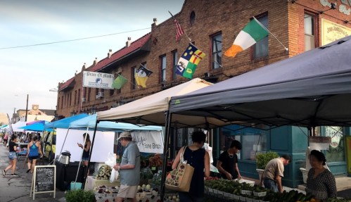 A bustling outdoor market with vendors under tents, flags flying, and shoppers browsing fresh produce and goods.