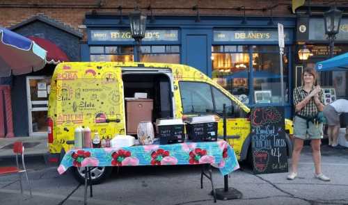 A colorful food truck with a table of drinks and snacks, set up on a street with a person standing nearby.